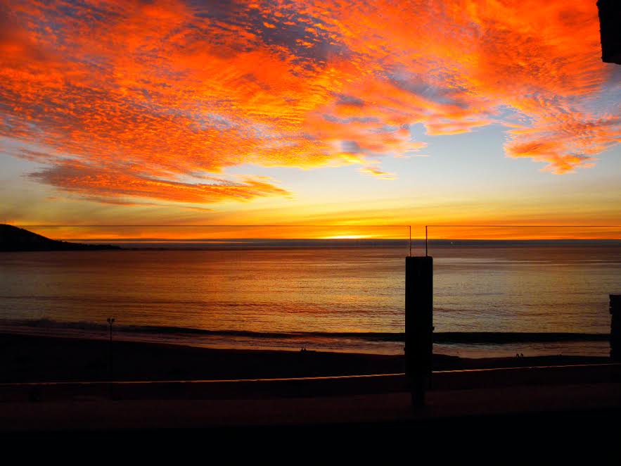 A stunning sunset over the ocean creates a vibrant palette of oranges and reds in the sky. The calm waters reflect the colors, while the horizon remains visible, creating a serene atmosphere. The scene is framed by a balcony railing, enhancing the open view.