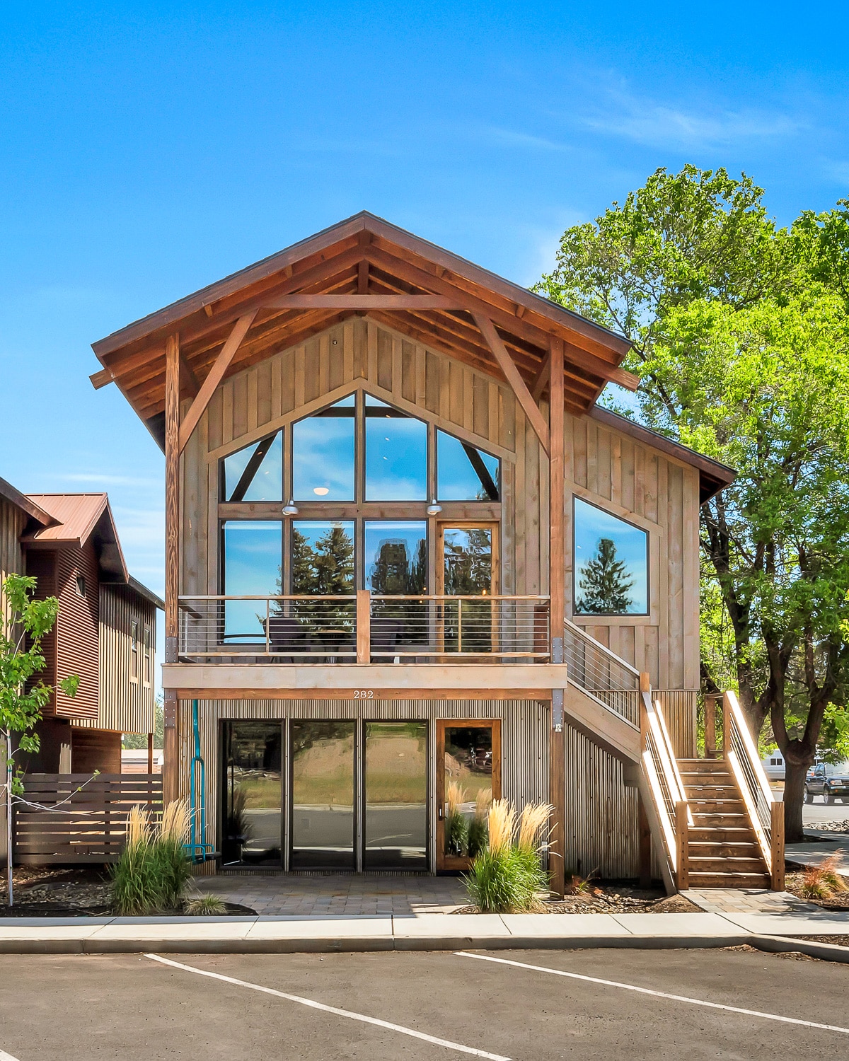 A modern wooden structure features a prominent peaked roof and large glass windows, allowing natural light to fill the interior. A welcoming entrance is framed by a spacious deck and surrounded by greenery, creating an inviting approach to the upper-level apartment.