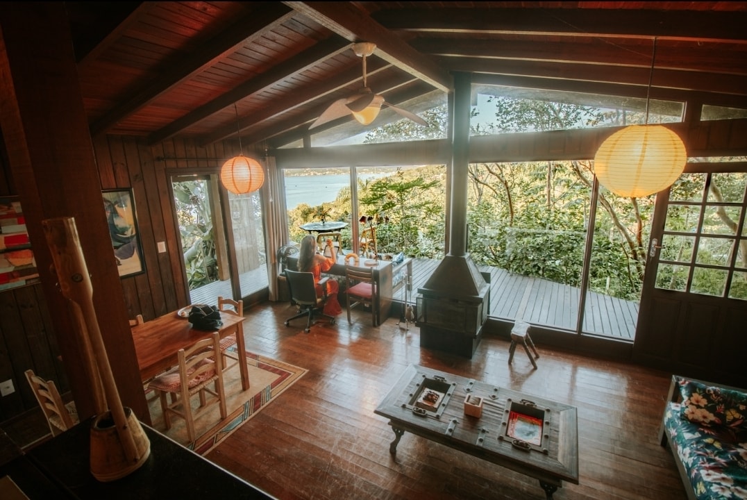 A spacious living area features large glass doors that invite natural light and views of greenery. A wooden dining table is positioned beside a cozy seating area, which is centered around a rustic coffee table and complemented by a wood-burning stove. Paper lanterns add a soft glow.