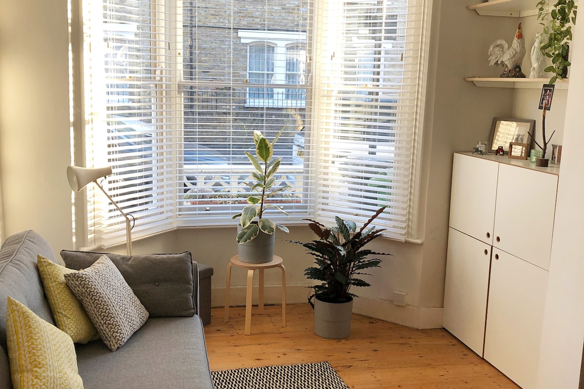 A light-filled living area features large bay windows adorned with white blinds. A cozy grey sofa with patterned cushions complements a wooden floor. A potted plant sits on a small table, while a low white cabinet displays decorative items, enhancing the space's minimalist style.