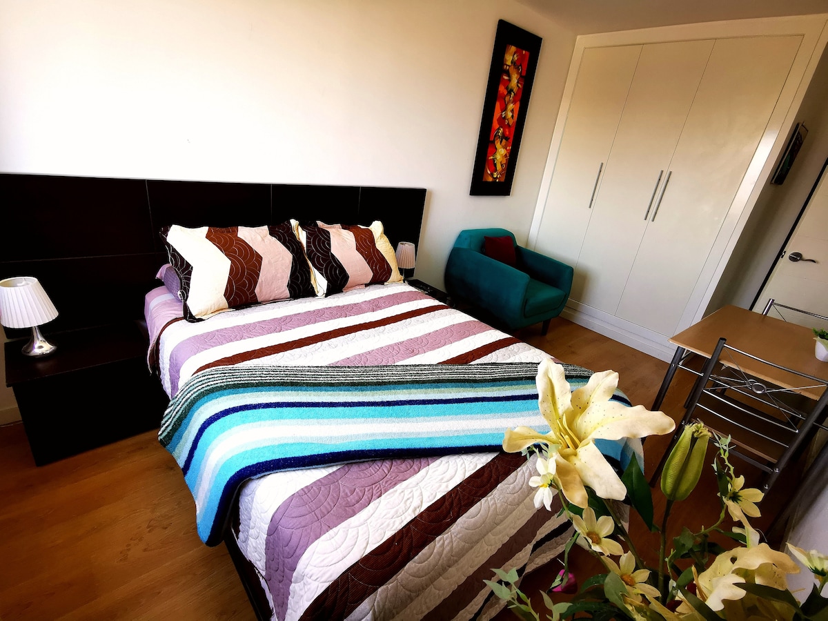 A bedroom features a spacious bed with decorative pillows and a patterned bedspread. A teal chair is positioned nearby, alongside a bedside lamp. A closet with sliding doors occupies one wall, while a small table adds functionality to the space. Floral decorations enhance the overall ambiance.