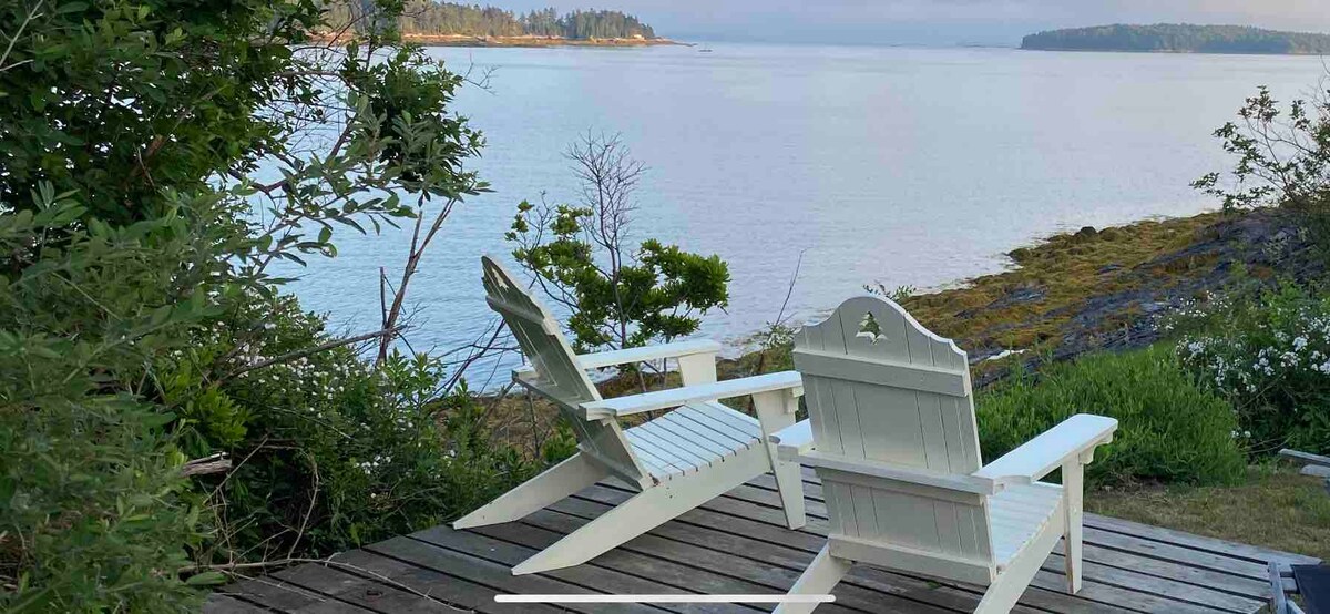 Two white Adirondack chairs are positioned on a wooden deck, facing tranquil waters of Mere Point Bay. Lush greenery surrounds the seating area, enhancing the natural scenery. The calm bay reflects soft morning light, while distant tree-lined shores are visible on the horizon.