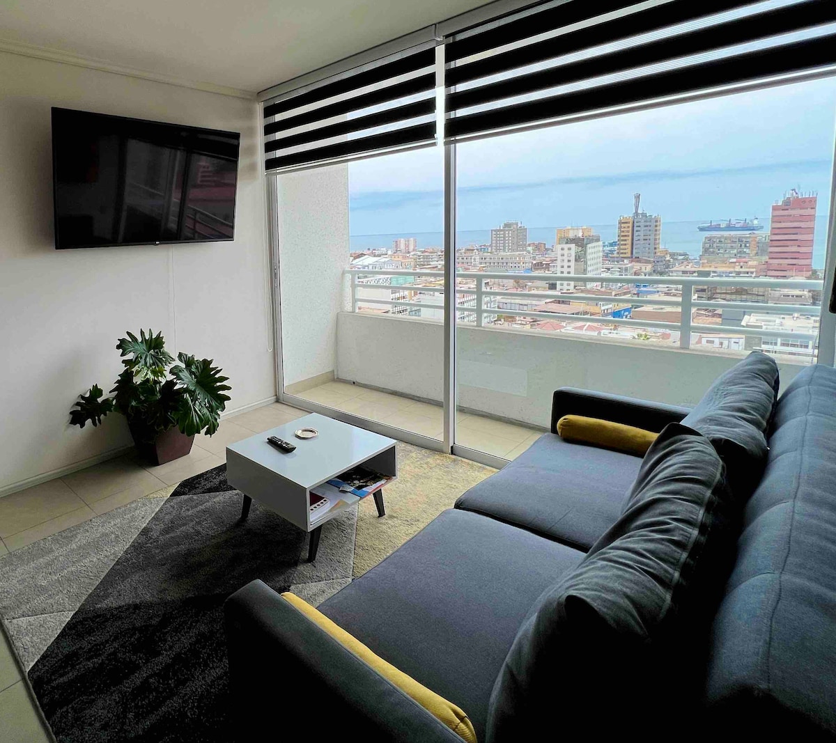 A spacious living area is viewed, featuring a large window with a panoramic sea view. A modern sofa with yellow accents is positioned next to a minimalist coffee table. A flat-screen TV is mounted on the wall, and a potted plant adds greenery to the space.