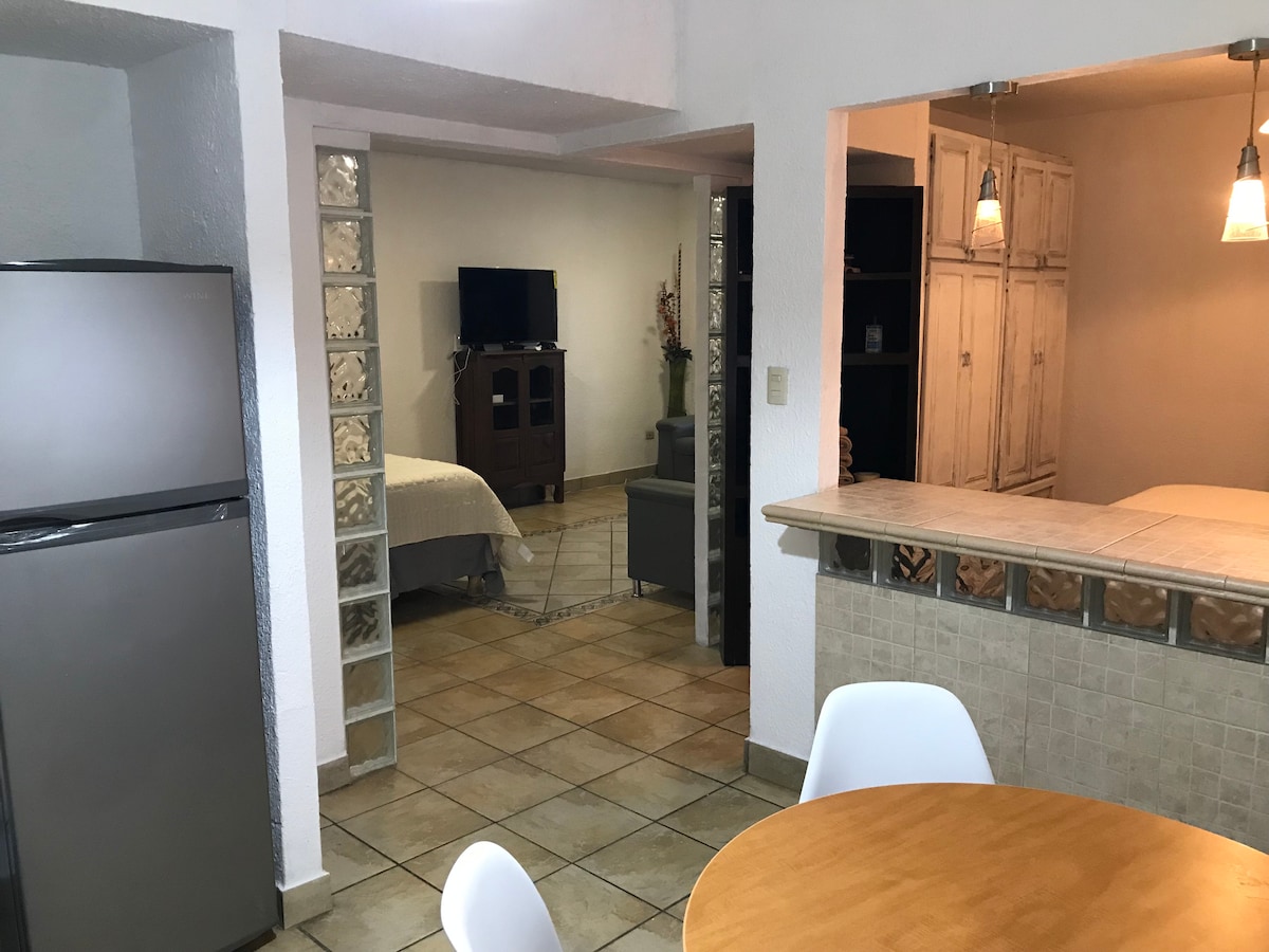 An open layout is presented, featuring a kitchenette that transitions to a dining area with a round table and white chairs. A living area with a television is visible, along with a cozy sleeping area, all accented by light-colored walls and tiled flooring.