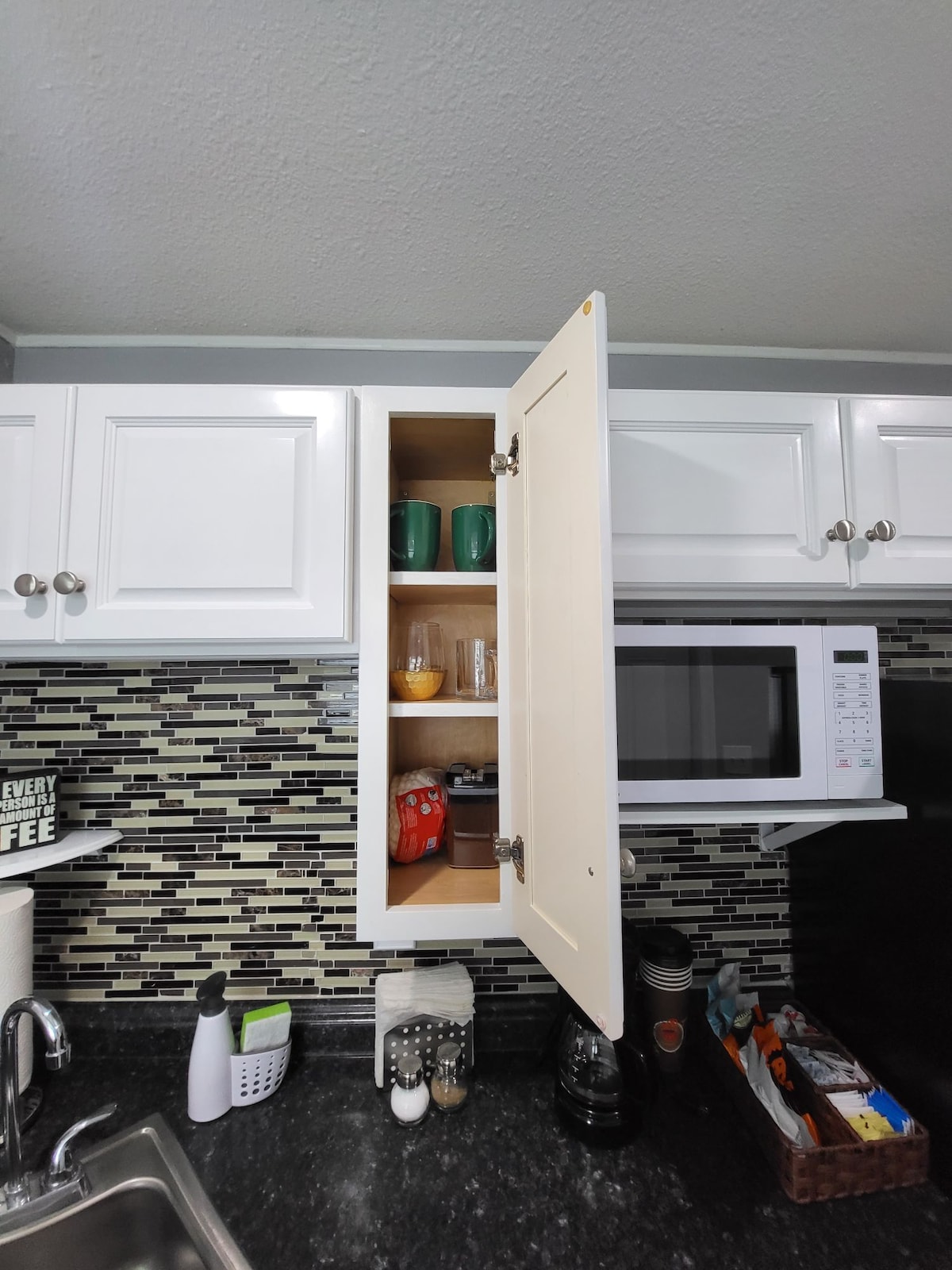 A white kitchen cabinet is shown with its door open, revealing a range of items including green mugs, a glass, and snacks. A microwave and various kitchen tools are visible on the countertop below, contributing to a functional workspace.
