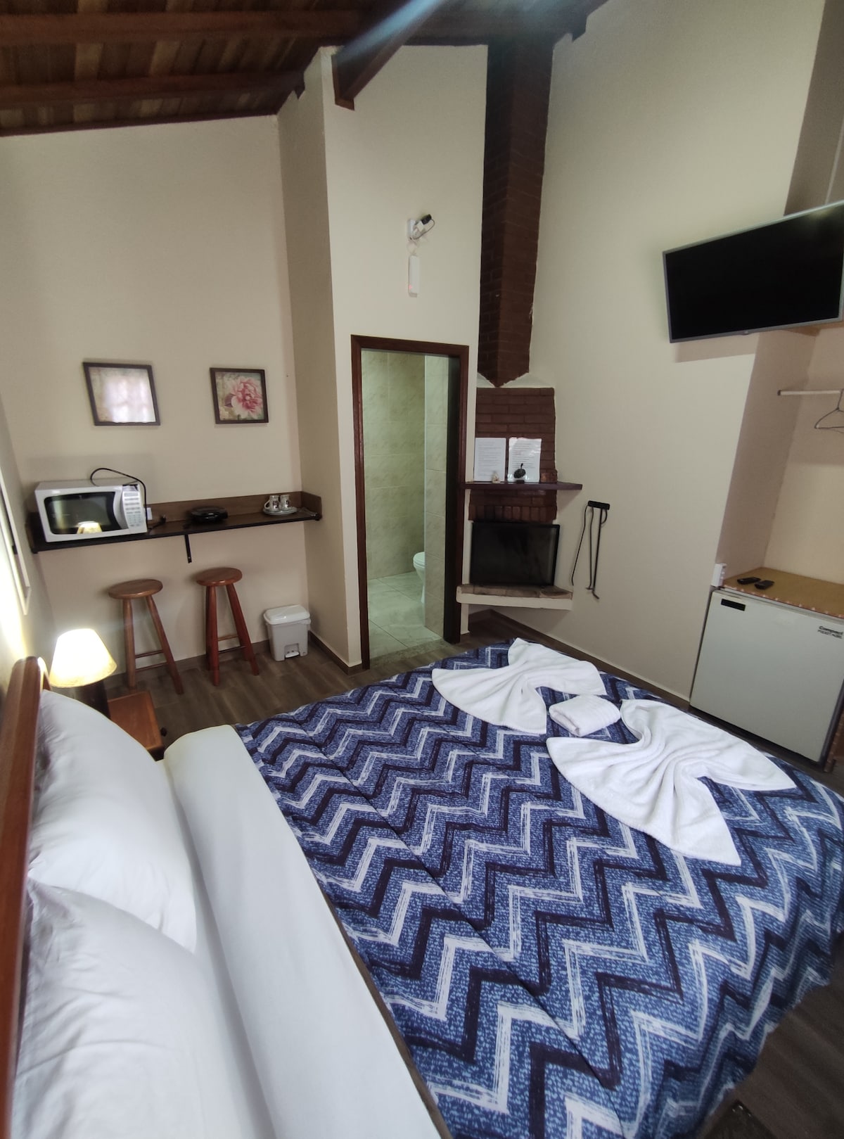 A warm sleeping area features a spacious bed with patterned bedding. A small kitchenette is seen, alongside a wooden shelf with decorative elements. A modern TV is mounted on the wall, and a bathroom doorway is visible. Light enters through a nearby window, highlighting the cozy ambiance.