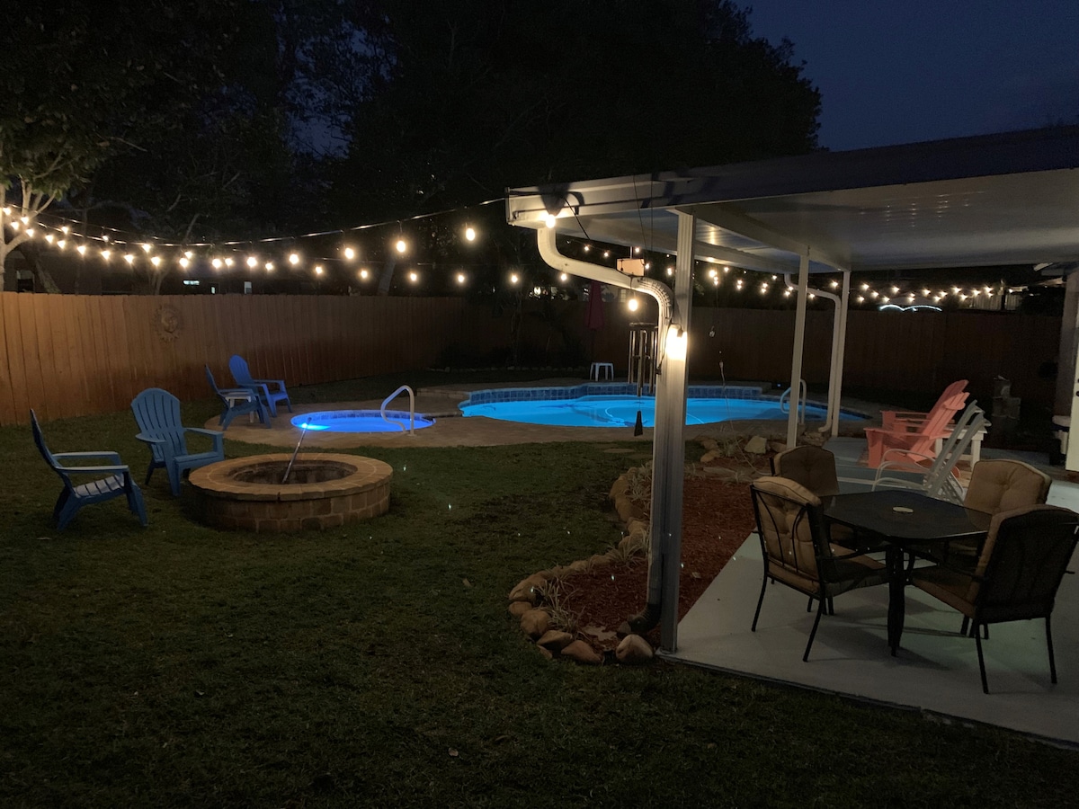 An outdoor area is illuminated by string lights, showcasing a pool with blue waters alongside a hot tub. Seating options include colorful Adirondack chairs and a patio table with four chairs. A fire pit is situated within a landscaped space, enhancing the night atmosphere.