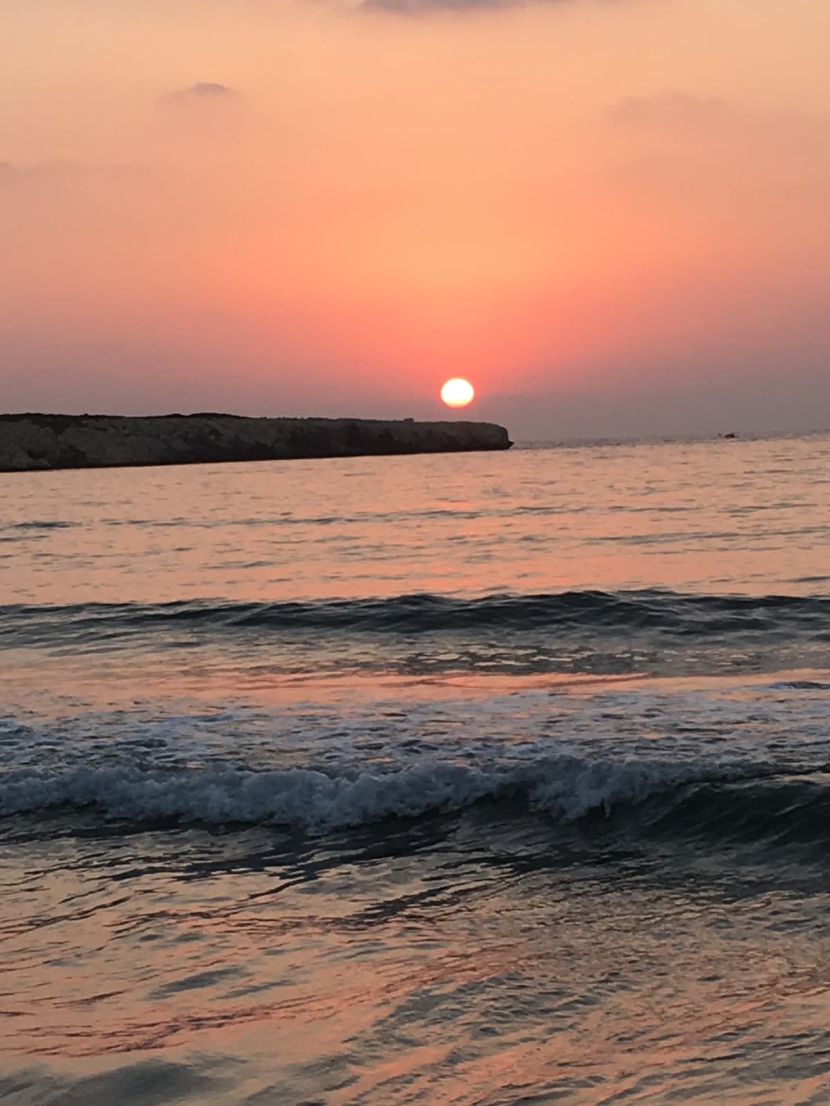 A serene sunset is depicted over the tranquil sea, with the sun partially descending behind a distant shoreline. Gentle waves softly lap at the beach, reflecting hues of orange and pink across the water, creating a calming atmosphere.