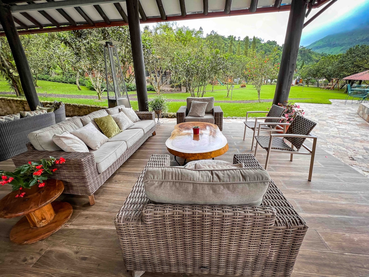 An outdoor seating area is furnished with a comfortable sectional sofa adorned with soft cushions and accompanied by a wooden coffee table. Two metal chairs add to the seating arrangement, with potted flowers placed nearby. Lush greenery and distant trees create a serene backdrop.
