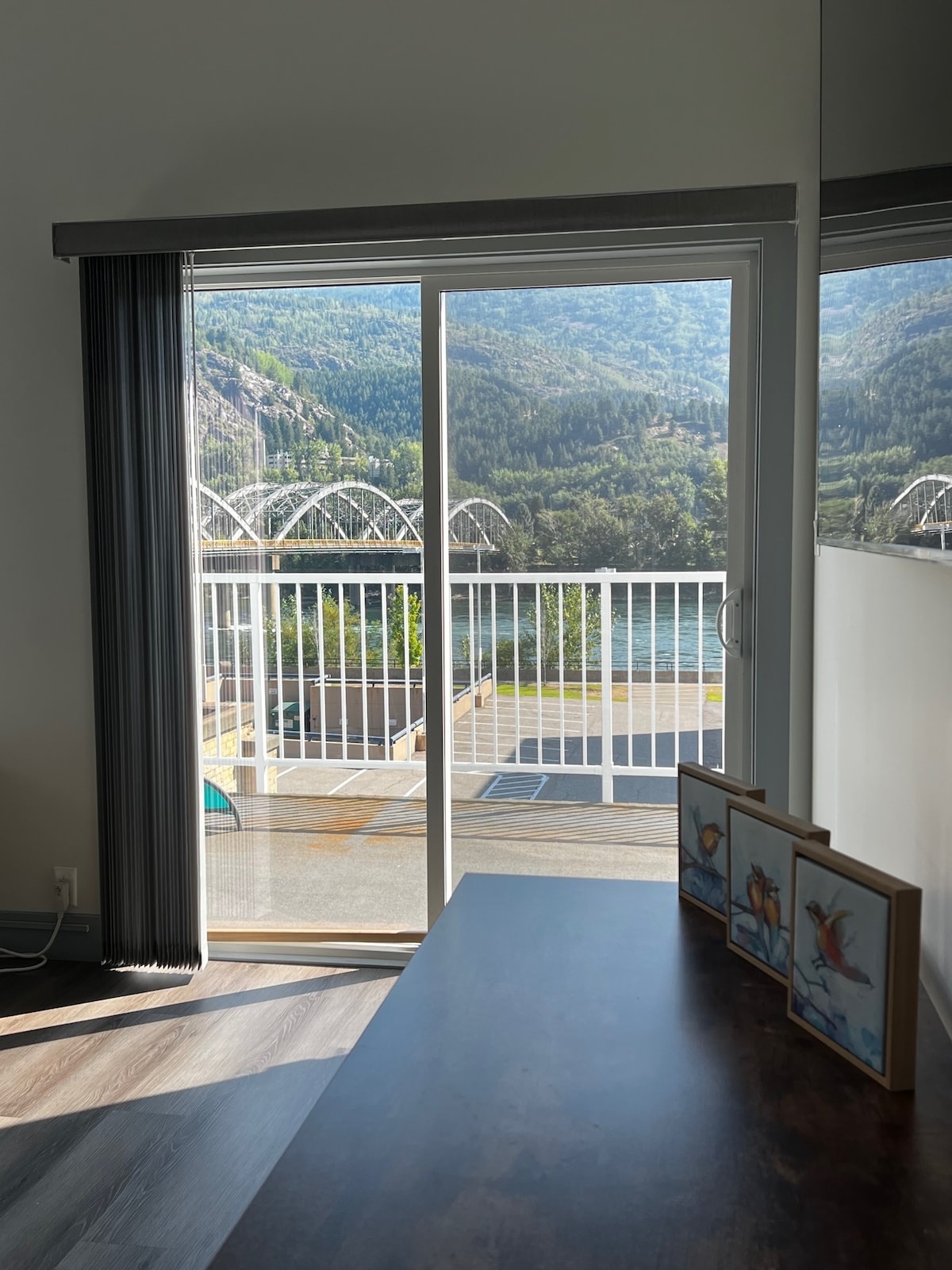 A glass sliding door opens to a balcony, offering captivating views of the surrounding mountains. A small table with framed artwork is positioned in the foreground, while the spacious outdoor area is visible beyond the railing.