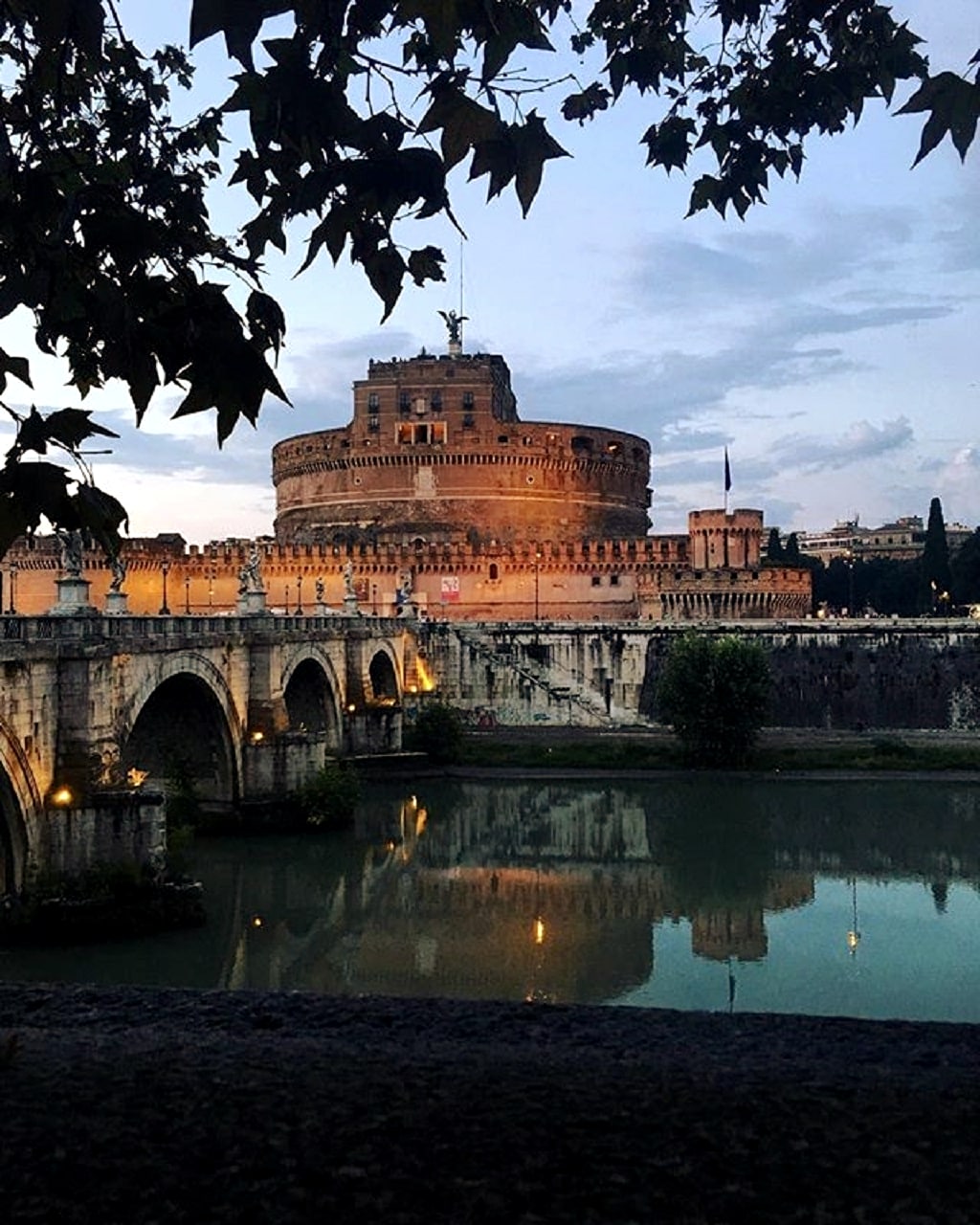 The Castel Sant'Angelo is illuminated at dusk, reflected in the calm waters of the Tiber River. Its historic architecture is framed by the surrounding greenery, with the soft glow of city lights casting a warm ambiance across the scene.