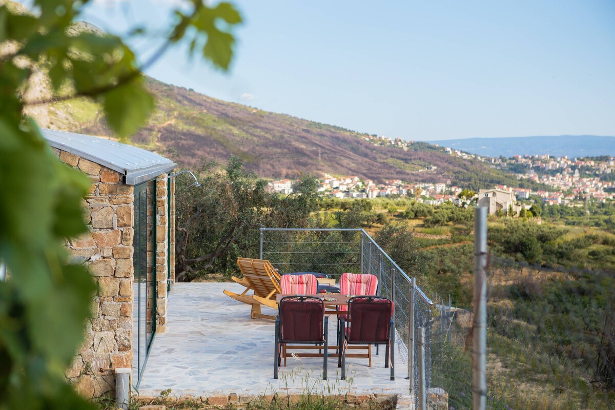 An outdoor terrace features a table surrounded by four chairs, with two sun loungers positioned nearby. A gentle slope of green hills extends in the background, with distant towns visible under a clear sky. The seating area is enclosed by a low fence.