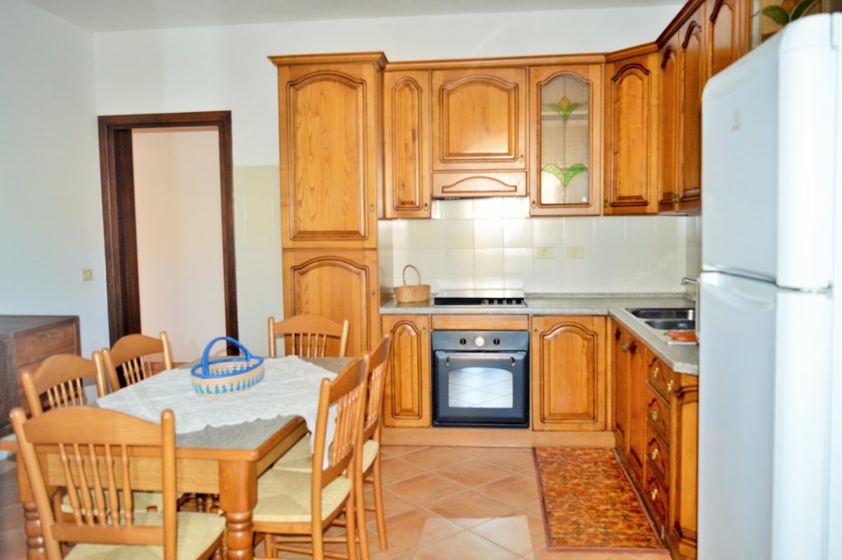 The kitchen features wooden cabinetry with a classic design and stone countertops. A dining table with ten wooden chairs occupies the center, while a white refrigerator is positioned against one wall. A stove and an oven are visible, providing essential cooking appliances.