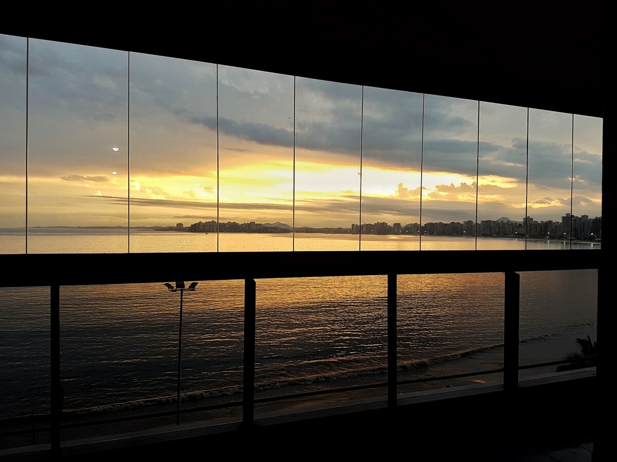 A panoramic view showcases a stunning sunset over the ocean, viewed through large glass windows. The horizon features a gradient of warm colors, reflecting on the water's surface. The calm beach is visible below, creating a serene atmosphere.