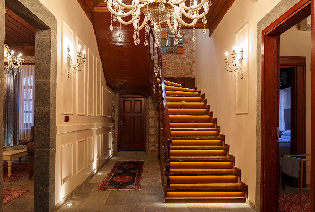 A well-lit entrance hall features a grand staircase with softly glowing steps. Elegant chandeliers hang from the ceiling, illuminating the space. Decorative wall panels create a classic ambiance, while plush seating can be seen in the adjacent area, offering a welcoming environment.