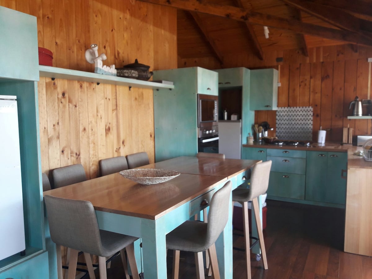 The kitchen is characterized by wooden walls and a high ceiling, featuring cabinetry in a soft blue hue. A spacious wooden dining table is surrounded by six stools. Kitchen appliances, including a refrigerator and cooking range, are visible, complemented by a well-organized countertop.