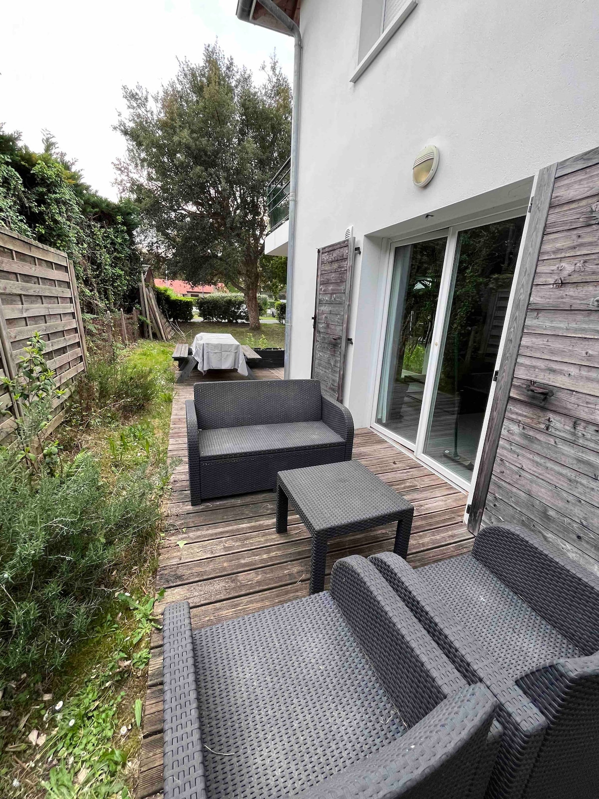 A spacious outdoor area is depicted featuring a wooden deck with comfortable seating arrangements. Dark wicker lounge chairs and a small table are positioned for relaxation. Vegetation surrounds the space, offering a green backdrop to the serene environment.