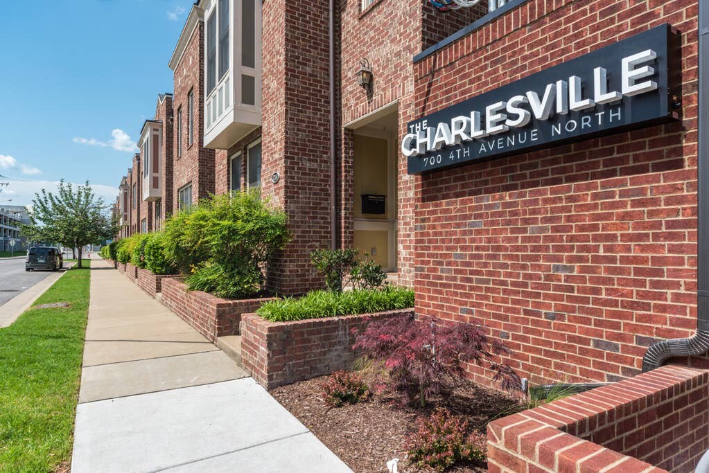 The exterior of The Charlesville exhibits a modern brick facade with large windows. A well-maintained sidewalk leads to the entrance, framed by neatly trimmed bushes and colorful landscaping. The surroundings showcase a paved pathway and the adjacent street, contributing to a welcoming urban environment.