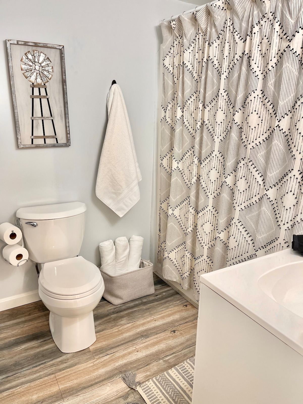 A clean and modern bathroom features a white toilet and a sleek sink with a minimalist design. Textured shower curtains display a geometric pattern, while neatly arranged towels are provided. A decorative wall piece adds character, and the flooring consists of wood-like tiles for a contemporary touch.