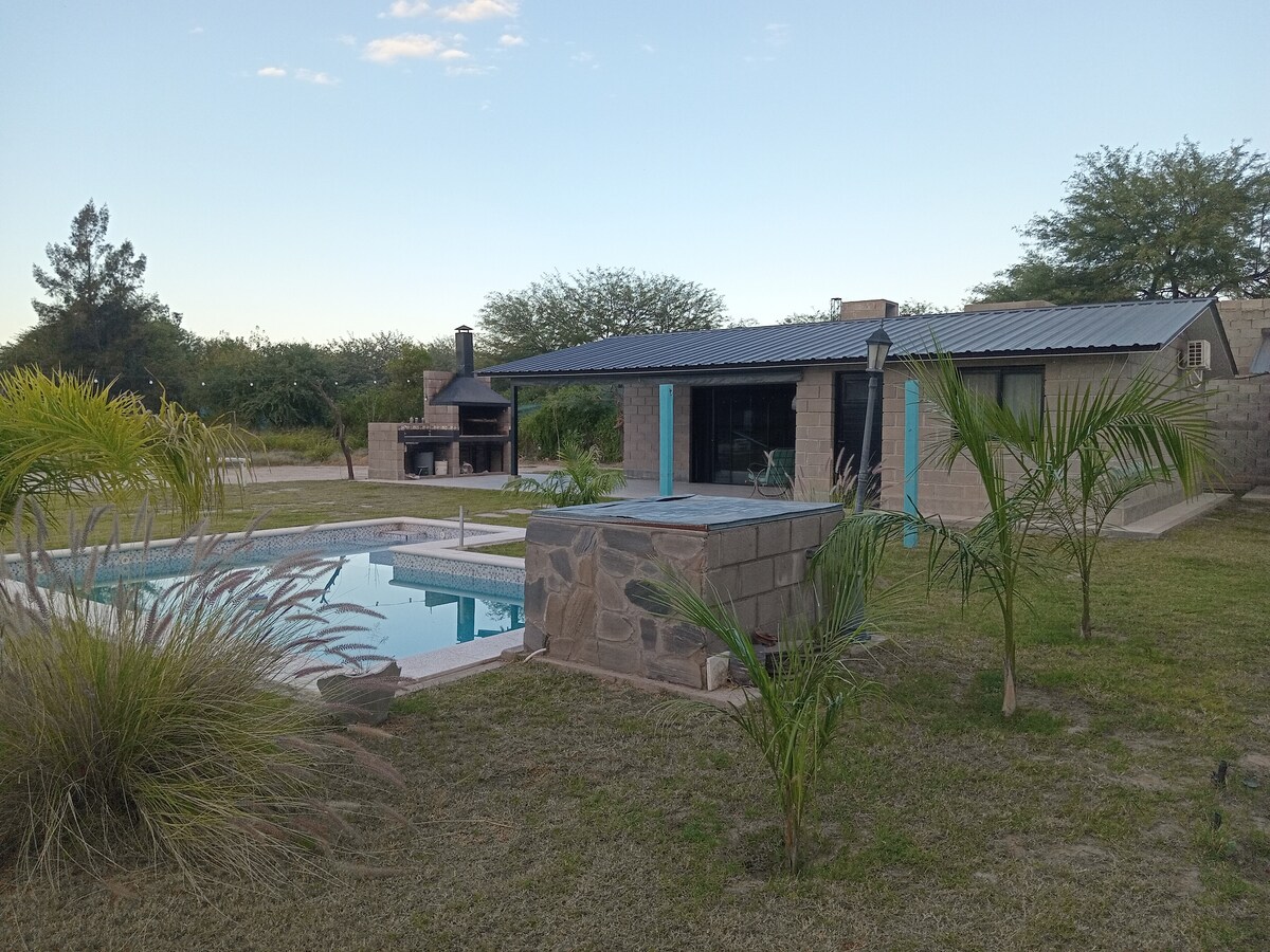 A tranquil outdoor space features a stone structure set against a clear sky. A swimming pool is situated nearby, complemented by greenery and decorative grasses. A barbecue area is visible, with a stone grill area adding to the inviting setting.