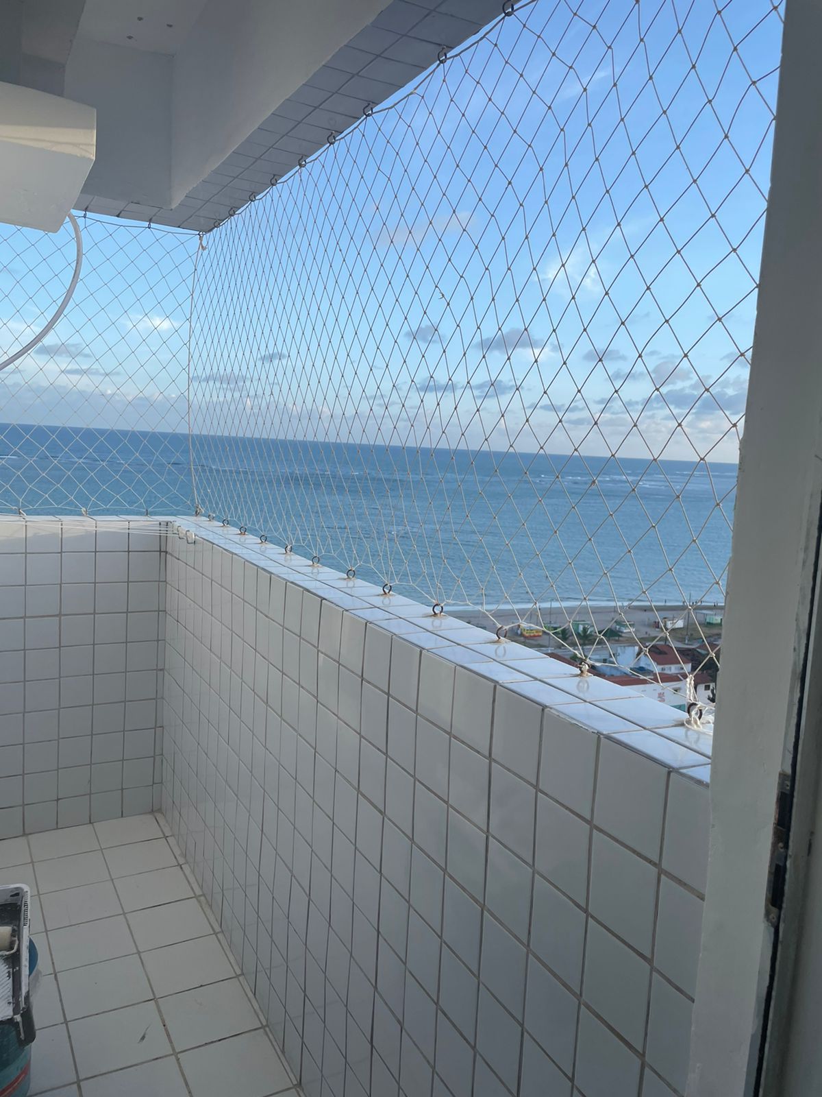 A serene ocean view is captured from the balcony, with a protective netting installed for safety. The tiled floor and white walls create a clean and bright environment, while the expansive sky and water offer a refreshing backdrop.