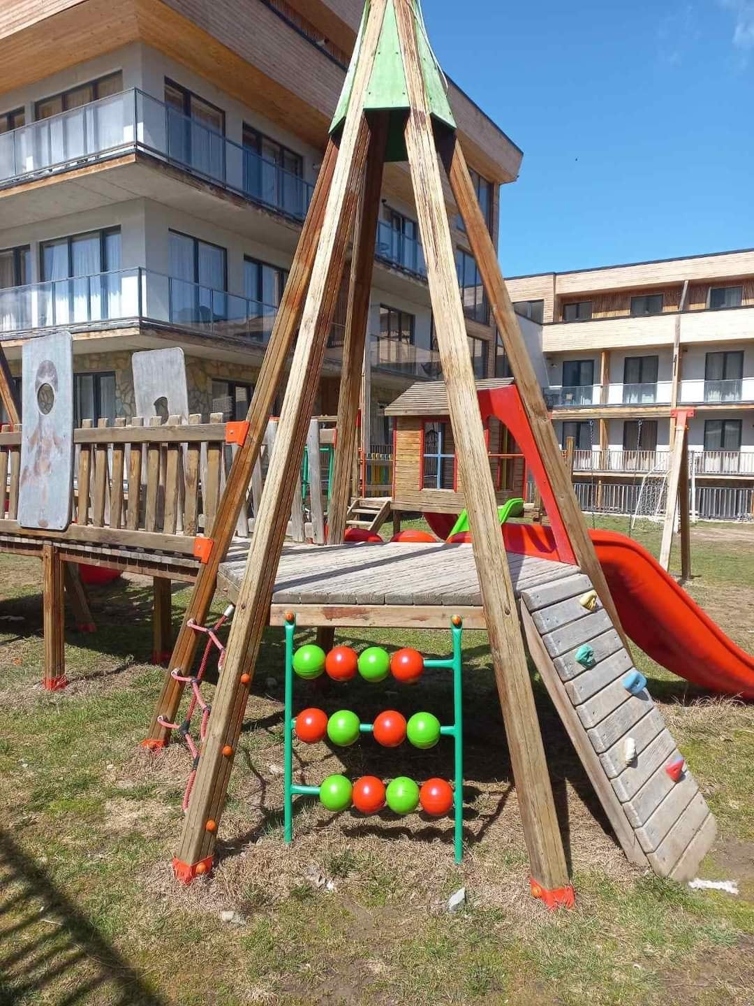 A wooden play structure is situated in a grassy area, featuring slides and climbing elements. Brightly colored panels are integrated into the design, alongside a play grid with red and green balls. The building in the background shows large balcony areas.