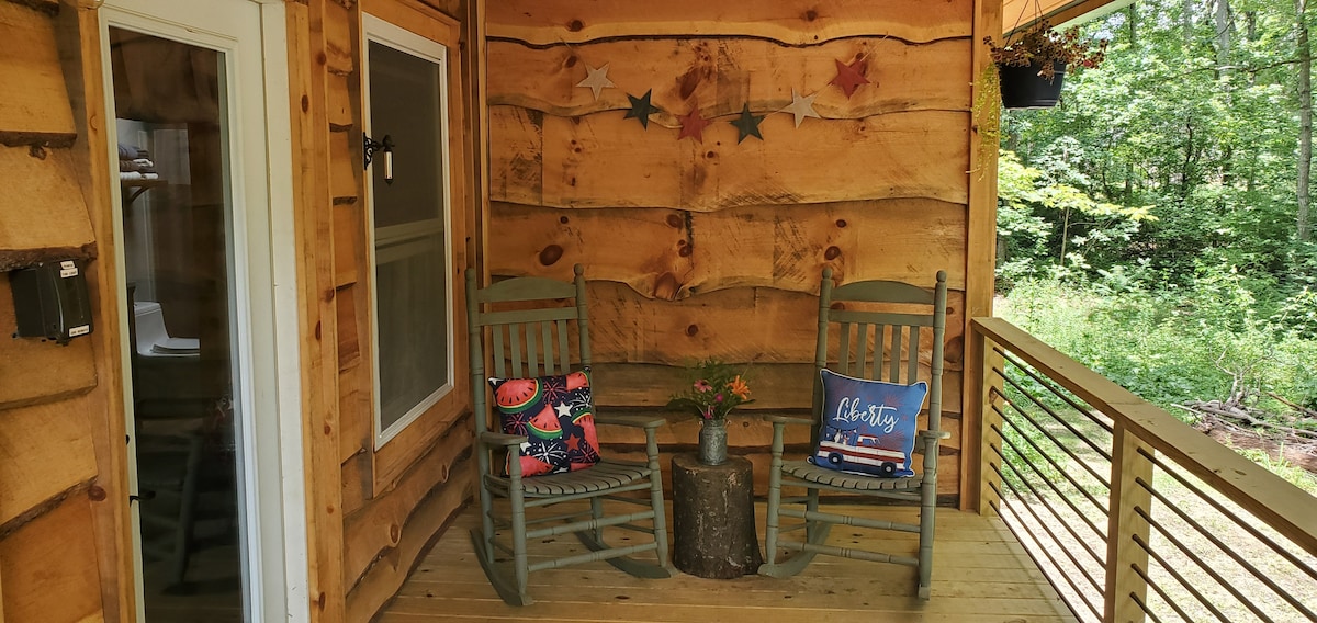 A serene porch area includes two rocking chairs, positioned around a rustic stump table. Decorative pillows add subtle color to the seating. The wooden walls exhibit a natural texture, while surrounding greenery enhances the peaceful ambiance.