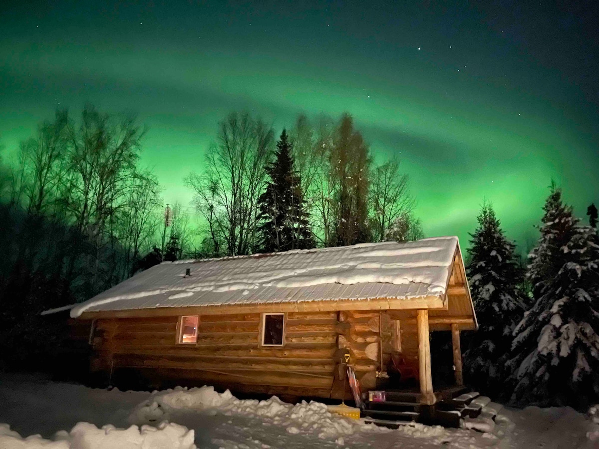 A rustic wooden cabin is illuminated under a vibrant green aurora borealis. The snow-covered roof and surrounding trees reflect the ethereal light, creating a serene winter scene. A peaceful and natural setting is offered, inviting connection with the Alaskan wilderness.