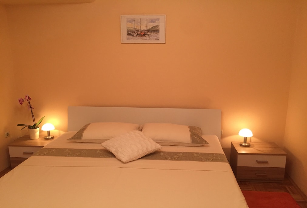A spacious bedroom is presented with a large bed featuring soft white linens and decorative pillows. Subtle lighting is provided by two bedside lamps on wooden nightstands. A small potted orchid adds a touch of greenery to the warm-toned wall, creating a calming environment.