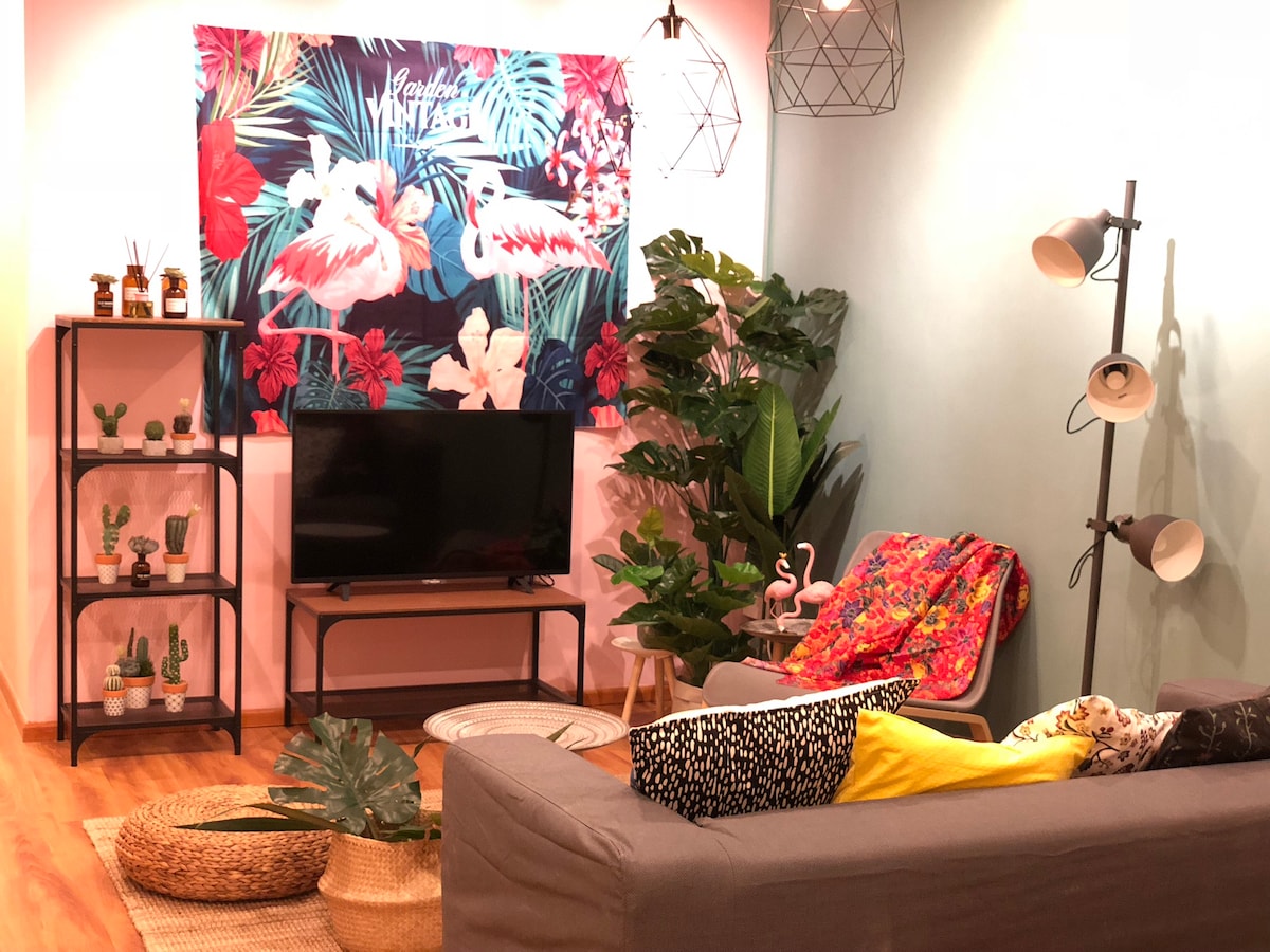 A welcoming shared lounge area is featured, embellished with a vibrant tropical backdrop. A spacious gray sofa is complemented by a yellow throw, while decorative plants and cacti are arranged throughout. A television is positioned on a modern stand, providing a comfortable space for relaxation.