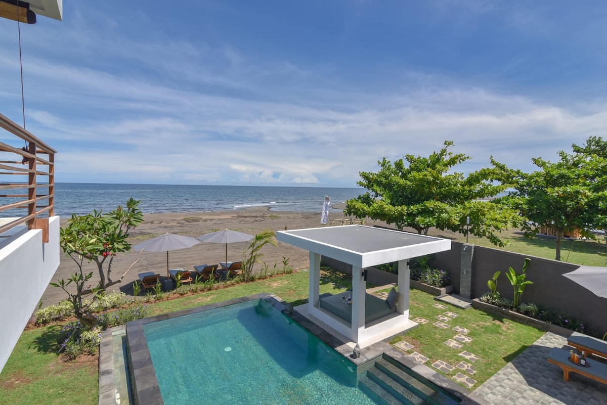 The image captures a private pool surrounded by lush greenery, leading to a beachfront view. Lounge chairs are positioned under umbrellas for relaxation. The serene ocean is visible in the background, meeting the clear blue sky, creating a peaceful coastal environment.
