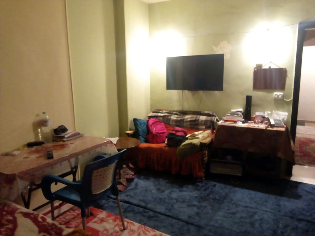 A cozy living area is furnished with sofa seating covered in patterned throws. A low table is surrounded by chairs, with various items and books displayed on its surface. A large television is mounted on the wall, and the room features soft lighting and muted wall colors.