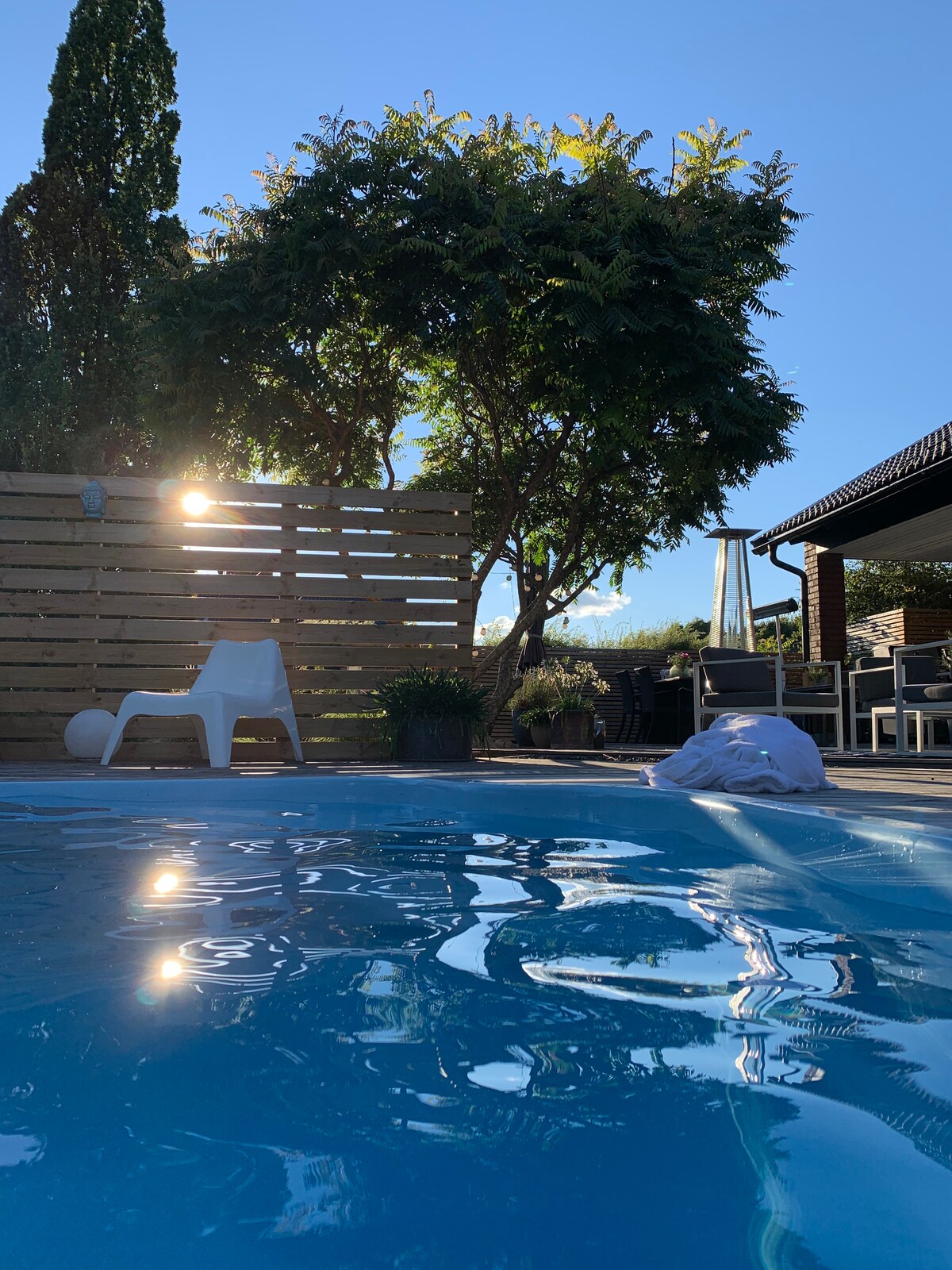 The image captures a serene outdoor scene featuring a clear swimming pool reflecting sunlight. A chair is positioned nearby, and a tree casts gentle shadows. A wooden fence partially encloses the space, enhancing the feeling of privacy and relaxation.