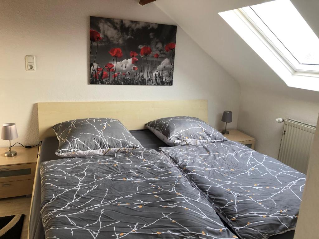 A spacious sleeping area features a large bed with two individual mattresses covered in patterned grey bedding. A light grey wall displays a vibrant red poppy artwork. Natural light enters through a skylight above, with a small lamp on each side of the bed and a wooden nightstand.