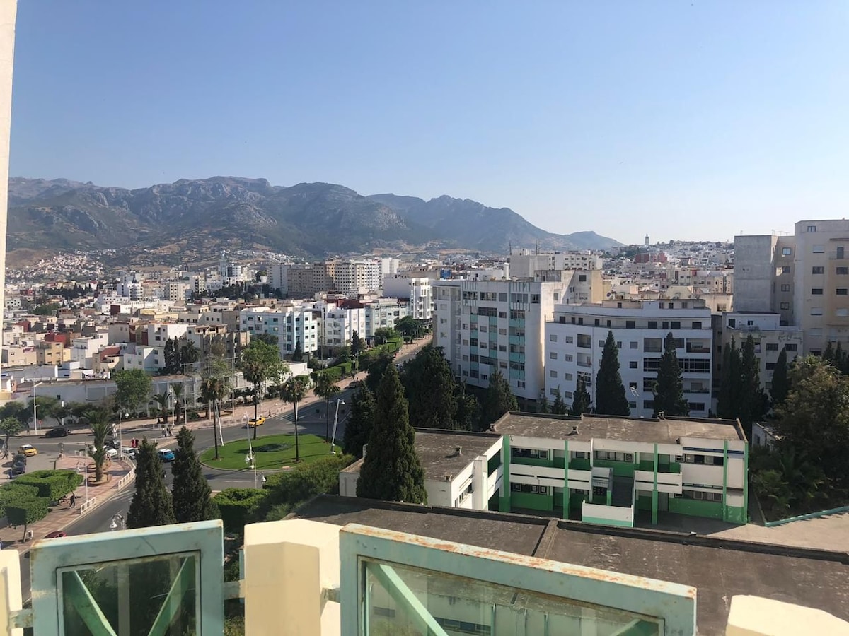 A panoramic view captures the surrounding cityscape and distant mountains. Buildings and greenery stretch across the scene, highlighting a vibrant urban landscape under a clear blue sky. The perspective includes residential complexes and tree-lined avenues.