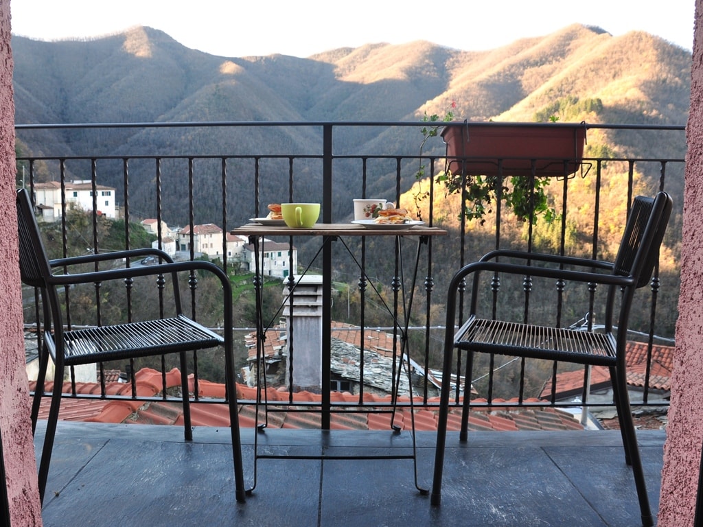 A balcony is shown with two chairs and a small table set for breakfast, featuring green and yellow cups and a plate of food. The view overlooks rolling mountains and a quaint village nestled in the valley below, surrounded by nature.