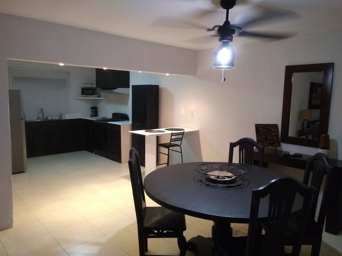An open-plan living space features a black dining table with four chairs, illuminated by a ceiling fan and light fixture. In the background, a kitchen is visible with modern appliances including a refrigerator, stove, and microwave, complemented by a breakfast bar with two stools.