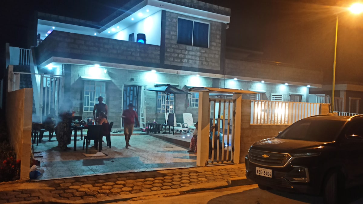 A well-lit exterior view showcases a two-story structure with a spacious patio. Outdoor furniture is arranged around a dining table, with individuals gathered, and the warm glow from blue lights highlights the entrance. A parked vehicle is visible in the foreground.