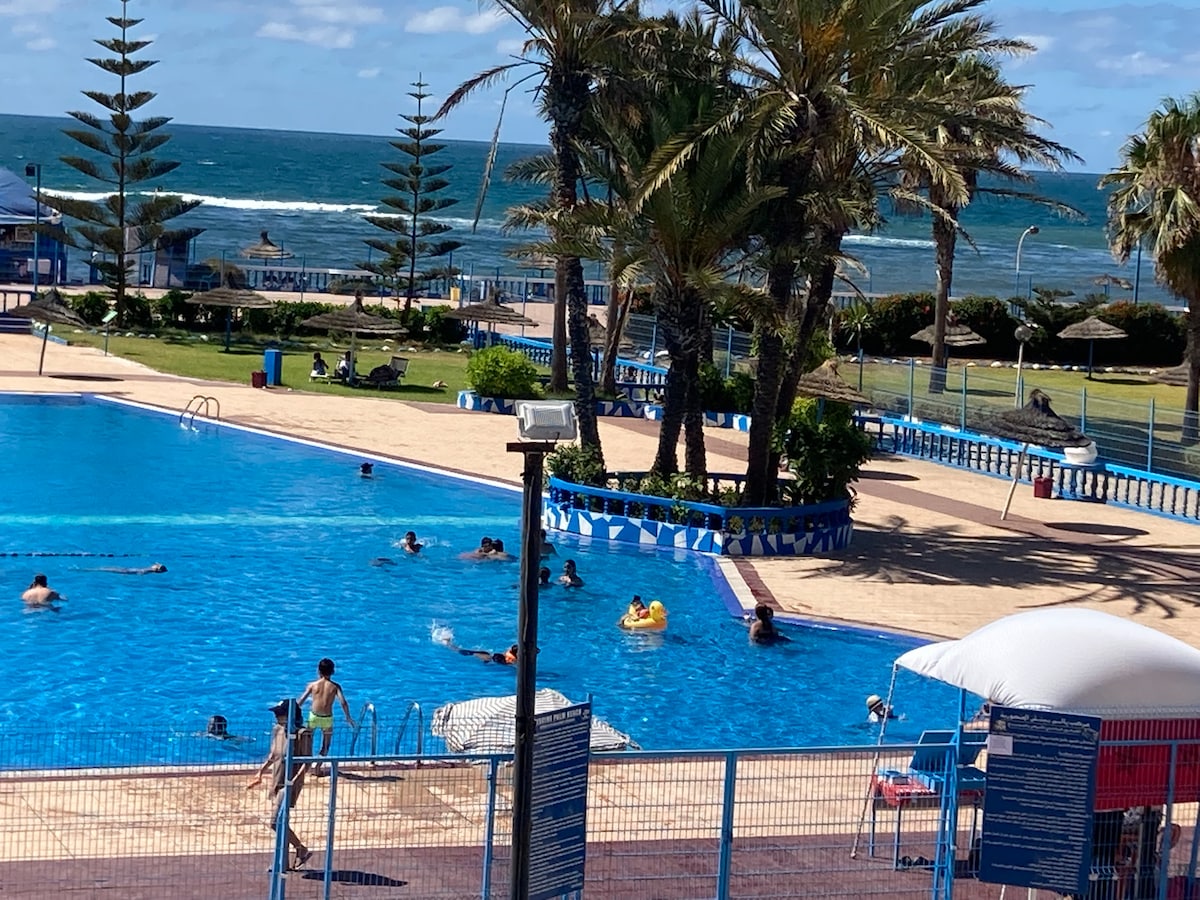 A large swimming pool is surrounded by sunlit areas and palm trees, with beachgoers enjoying the water. The ocean can be seen in the background, offering a scenic view. Loungers and umbrellas are positioned near the pool, creating a relaxed atmosphere.