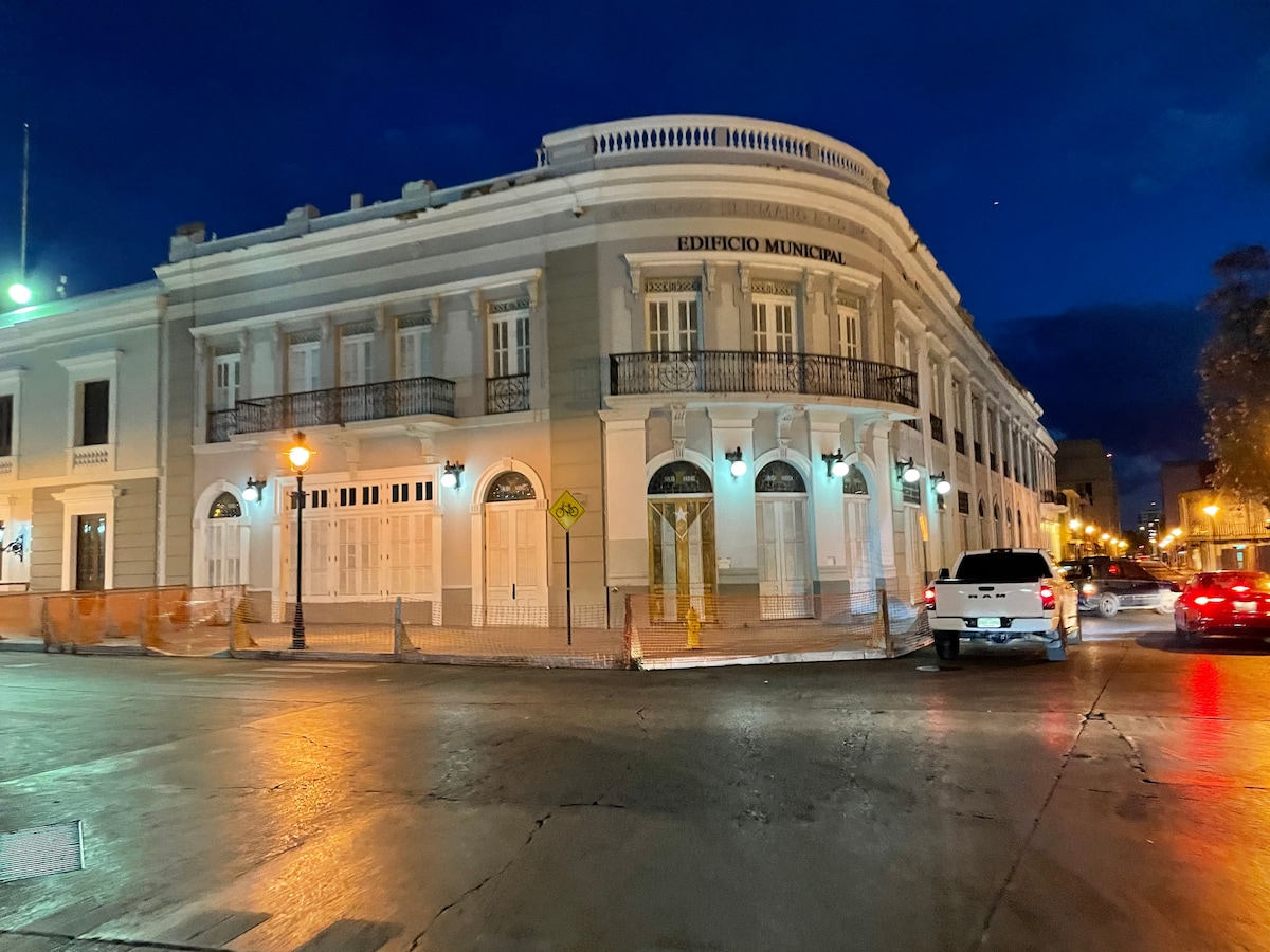 An elegant historic building is displayed under the evening sky, featuring a classic façade with balustrades and large windows. Streetlights cast a warm glow on the surrounding area, with vehicles parked along the curb, enhancing the ambiance of the neighboring streets.