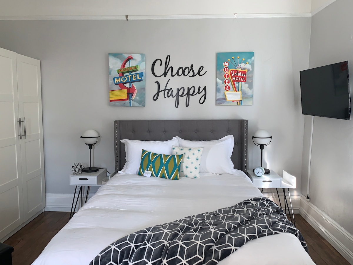 A spacious bedroom features a large bed with white linens and decorative pillows. Colorful artwork is displayed above the bed, complemented by sleek nightstands with modern lamps. A flat-screen TV is mounted on the wall, and a wardrobe is positioned nearby, enhancing the room's functionality.