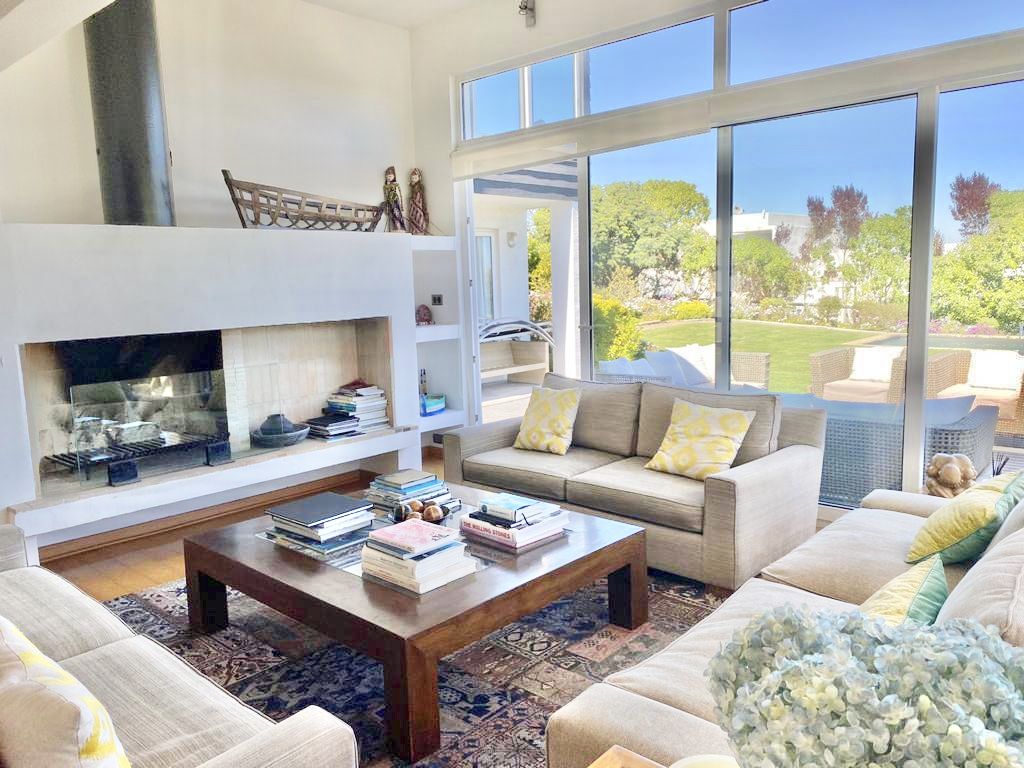 A bright living area features a modern design with large windows offering views of the garden. Two light-colored sofas are arranged around a dark wooden coffee table stacked with books. A fireplace stands against the wall, adding a cozy touch to the space.