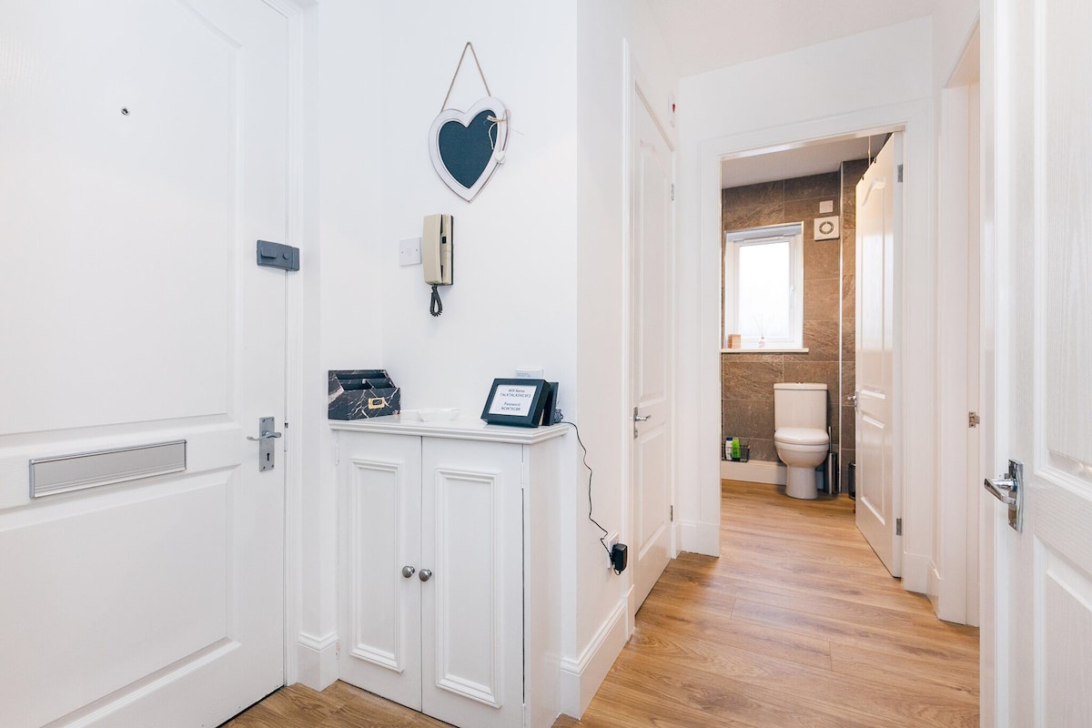 The entrance area showcases a clean layout with light-colored walls and wooden flooring. A heart-shaped wall decor piece adds a touch of charm. A side table holds informational materials, and a door in the background leads to a shower room featuring modern fixtures.