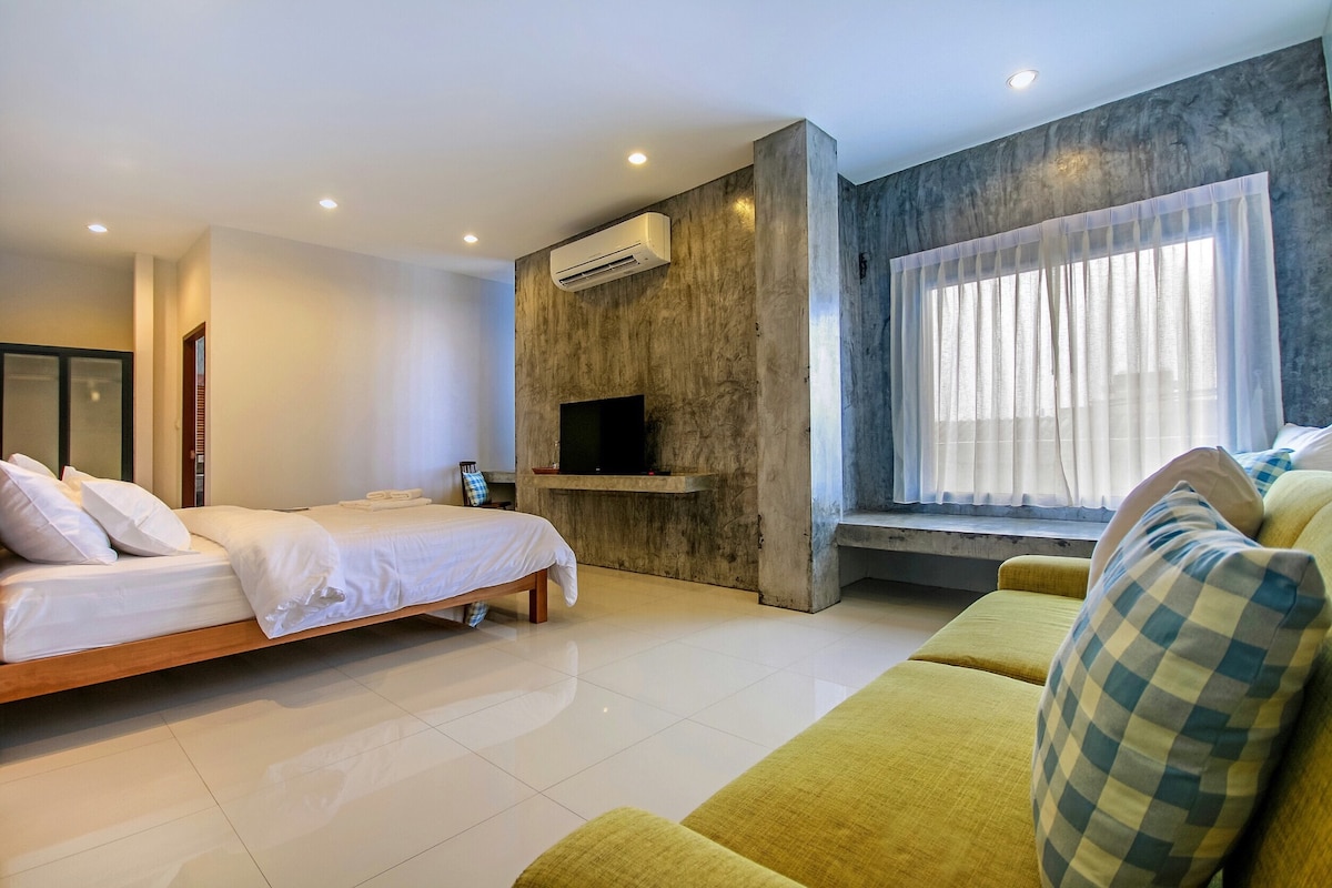 A spacious room is equipped with a large bed featuring white linens. A sleek television is mounted on a concrete wall, alongside a cozy seating area with a bright yellow couch and checkered cushions. Natural light enters through a large window dressed with sheer curtains.