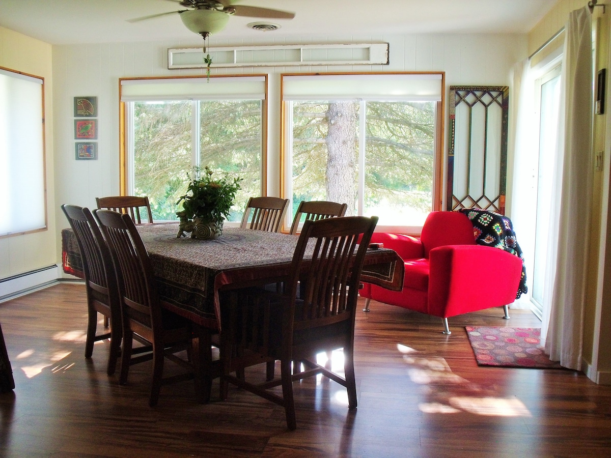 A spacious dining area features a large wooden table surrounded by eight chairs, bathed in natural light from expansive windows. A vibrant red armchair and nearby plants add a pop of color to the room, while bamboo flooring provides warmth.