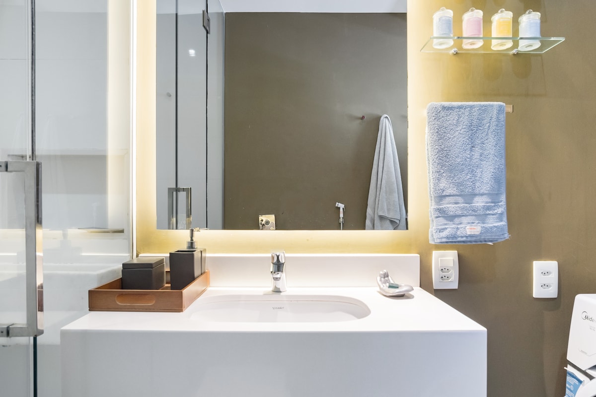 A modern bathroom features a sleek countertop with a stylish basin and an organized tray holding toiletries. Soft lighting reflects off the mirror, enhancing the space. A towel hangs neatly beside the sink, while colorful shampoo bottles are displayed on a glass shelf above.