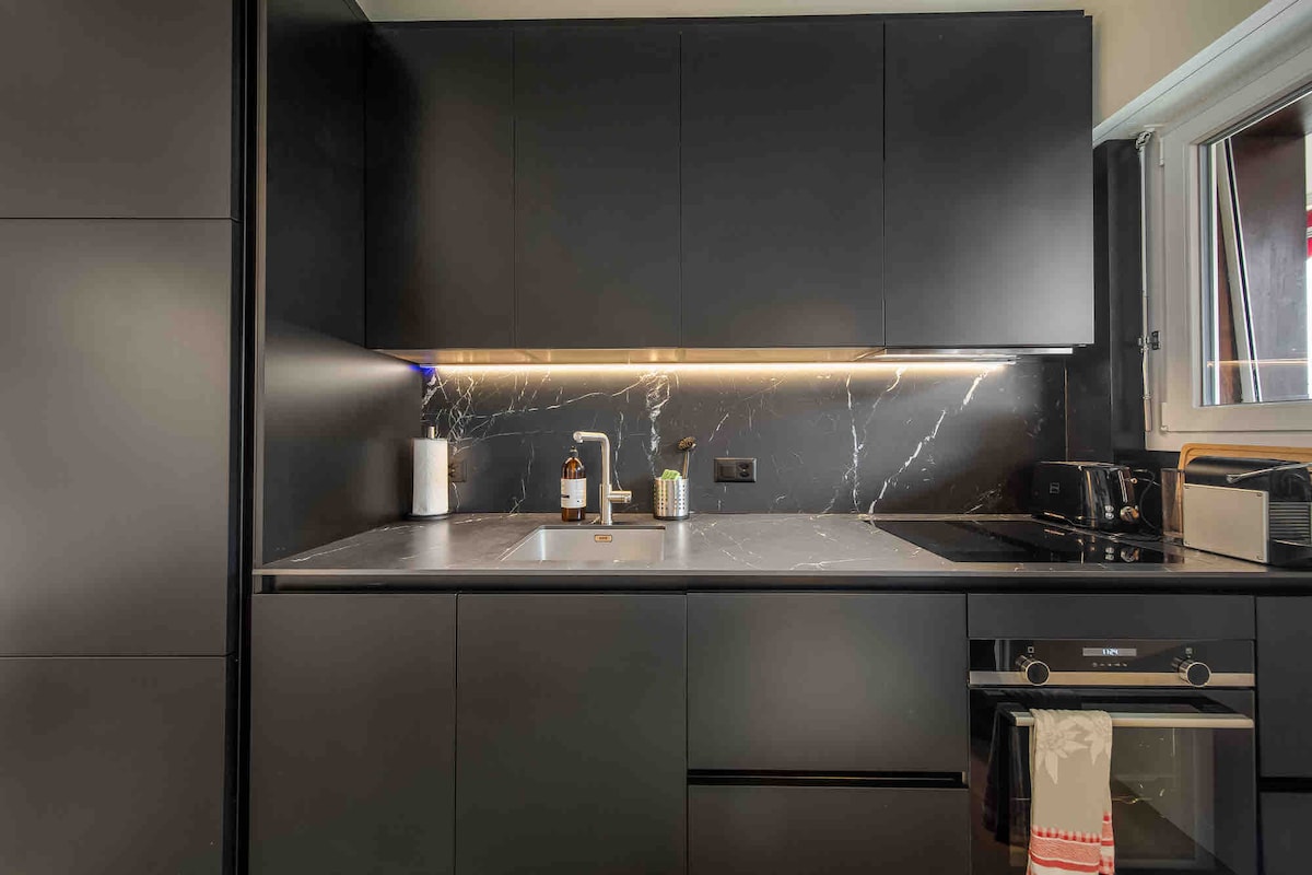 The modern kitchen features sleek black cabinetry with a marble backsplash. A stainless-steel sink is accompanied by a stylish faucet, and appliances include a toaster and coffee maker. Under-cabinet lighting highlights the countertop, providing a functional space for meal preparation.
