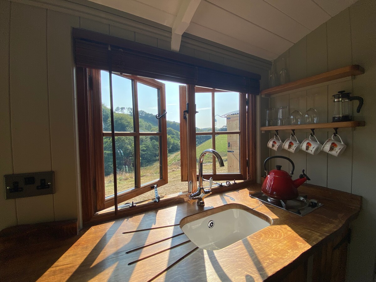 A rustic kitchen space features a wooden countertop with a sink and a gas cooker. Natural light floods the area through an open window adorned with multiple panes, overlooking verdant fields. Above, a shelf holds glassware and a red kettle, enhancing the functional charm.
