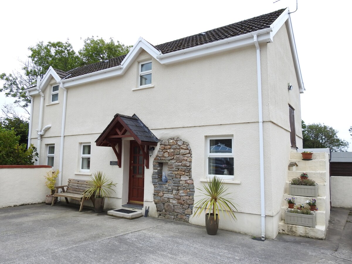 The exterior of the cottage features a simple yet elegant entrance with a stone accent wall and a wooden porch. Flanking the entrance are planters with greenery. A spacious driveway leads to the home, which has multiple windows allowing natural light.