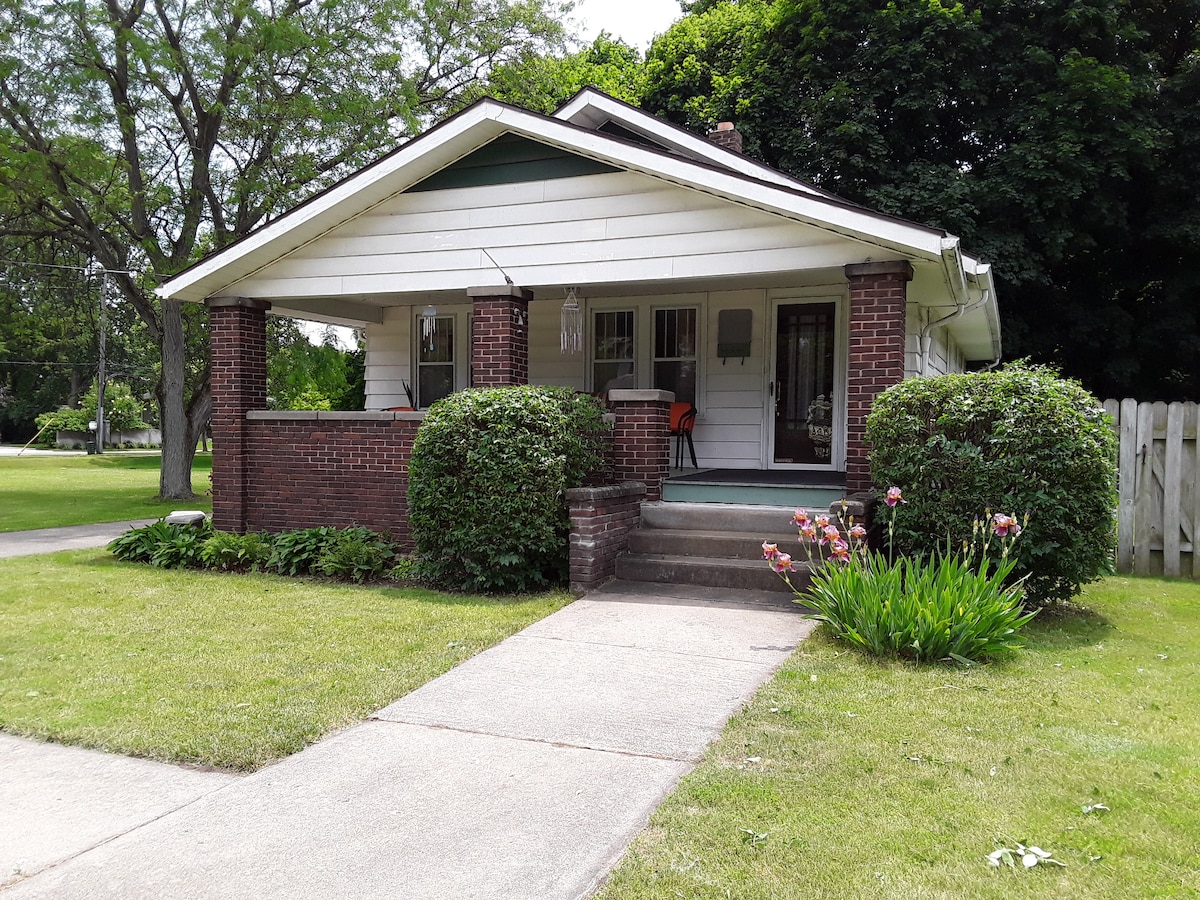 The front of the home showcases a welcoming porch supported by brick columns. Green shrubs and seasonal flowers line the entrance, while a paved path leads to the front steps. Lush grass surrounds the home, emphasizing the inviting, safe atmosphere of the neighborhood.